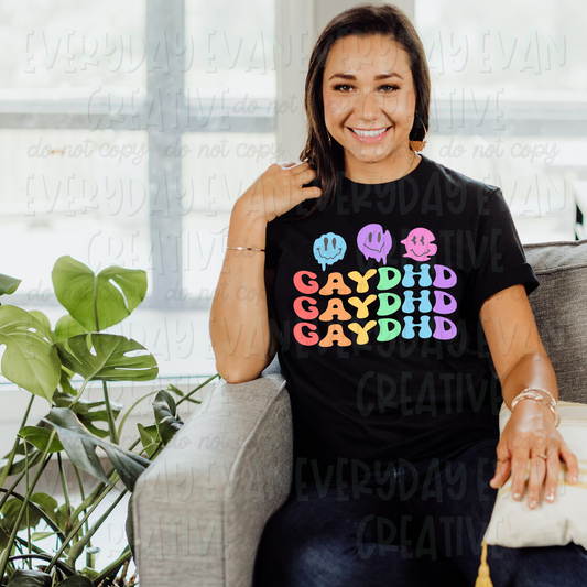A brunette white woman sits on a couch modeling a black t-shirt. The design on the shirt has three retro melted smiley faces in blue, purple and pink. Underneath in a wavy retro stacked pastel rainbow font it says "GAYDHD" as a play on ADHD.