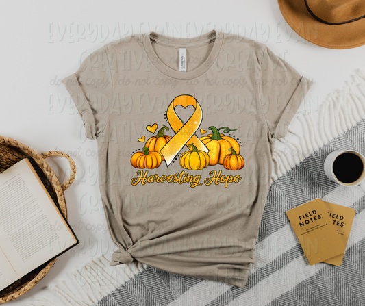 Image displays a t-shirt surrounded by miscellaneous desk items. The shirt's image includes several yellow pumpkins with green steps, a large yellow ribbon in the middle of the pumpkins, and small yellow hearts and black dots surrounding the design. The ribbon has a heart for the middle loop. Under the image, there is yellow script text that reads, "Harvesting Hope"