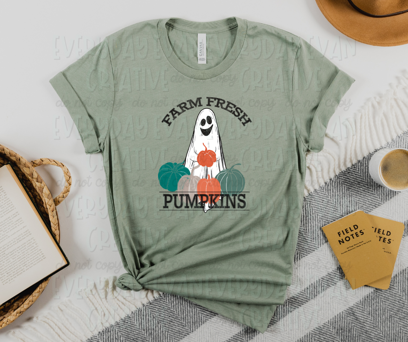 Image displays a t-shirt with miscellaneous desk items surrounding it. The shirt's image includes a smiling sheet ghost holding an orange pumpkin, several different colored pumpkins surrounding the bottom of it, and text that reads, "farm fresh" at the top and "pumpkins" at the bottom in serif font.  