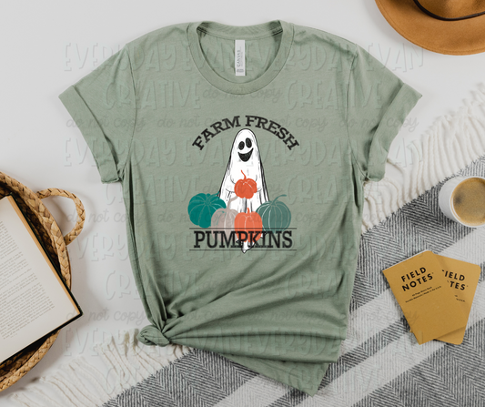 Image displays a t-shirt with miscellaneous desk items surrounding it. The shirt's image includes a smiling sheet ghost holding an orange pumpkin, several different colored pumpkins surrounding the bottom of it, and text that reads, "farm fresh" at the top and "pumpkins" at the bottom in serif font.  