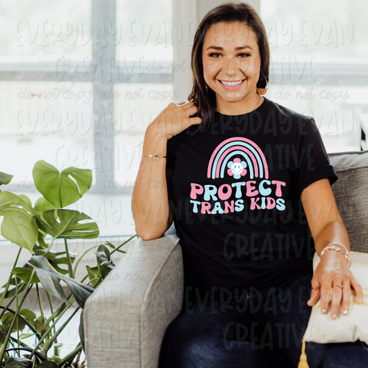 A brunette white woman sits on a couch modeling a black t-shirt. The design on the t-shirt has a pastel blue and pink rainbow with a blue, pink, and white smiley face groovy flower in the middle of it. Underneath in alternating pink and blue retro wavy font, it says "protect trans kids."