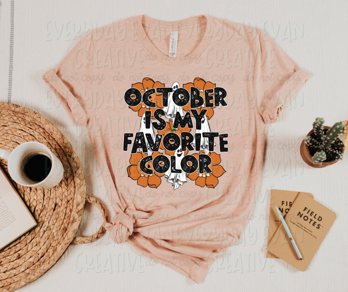 Image displays a t-shirt surrounded by miscellaneous desk items. The t-shirt is pink in color, and has a flower and sheet ghost design underneath creative, black, block text that reads, "October is my favorite color." The flowers and sheet ghosts alternate behind the text in three rows of three.