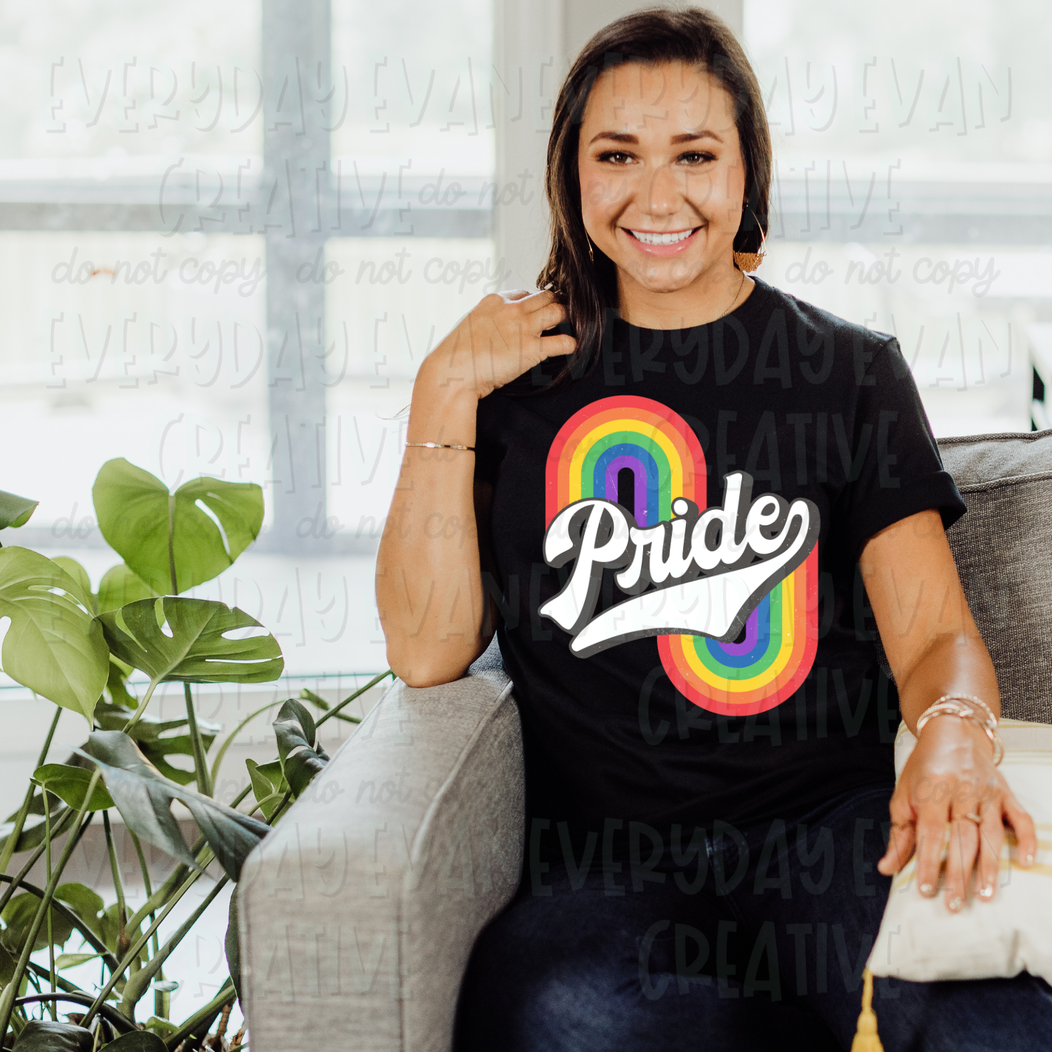 A brunette white woman sits on a couch modeling a black t-shirt. The design on the shirt centers the word "pride" in white with a gray shadow in a baseball team style font. There is a rainbow on both sides of the word. The left is rightside up and the right is upside down. 