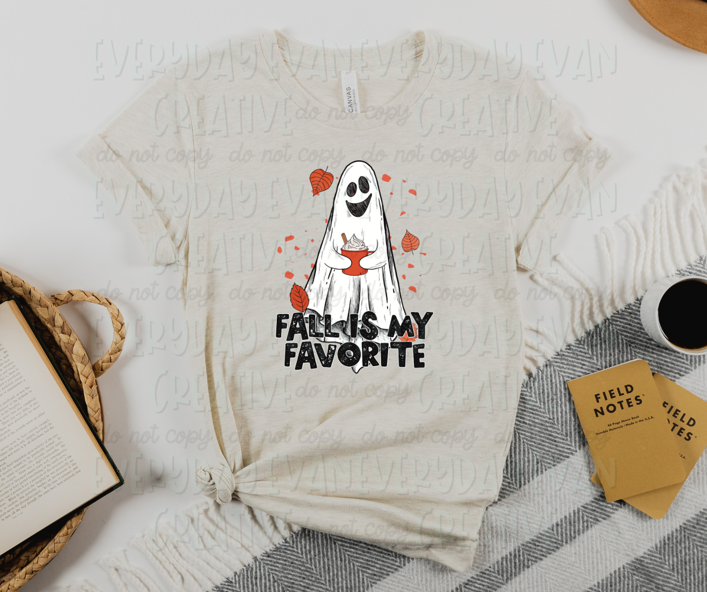 Image displays a t-shirt surrounded by miscellaneous desk items. The shirt's design has a smiling sheet ghost holding an orange mug surrounded by fall leaves. Under the sheet ghost, creative block text reads, "fall is my favorite."