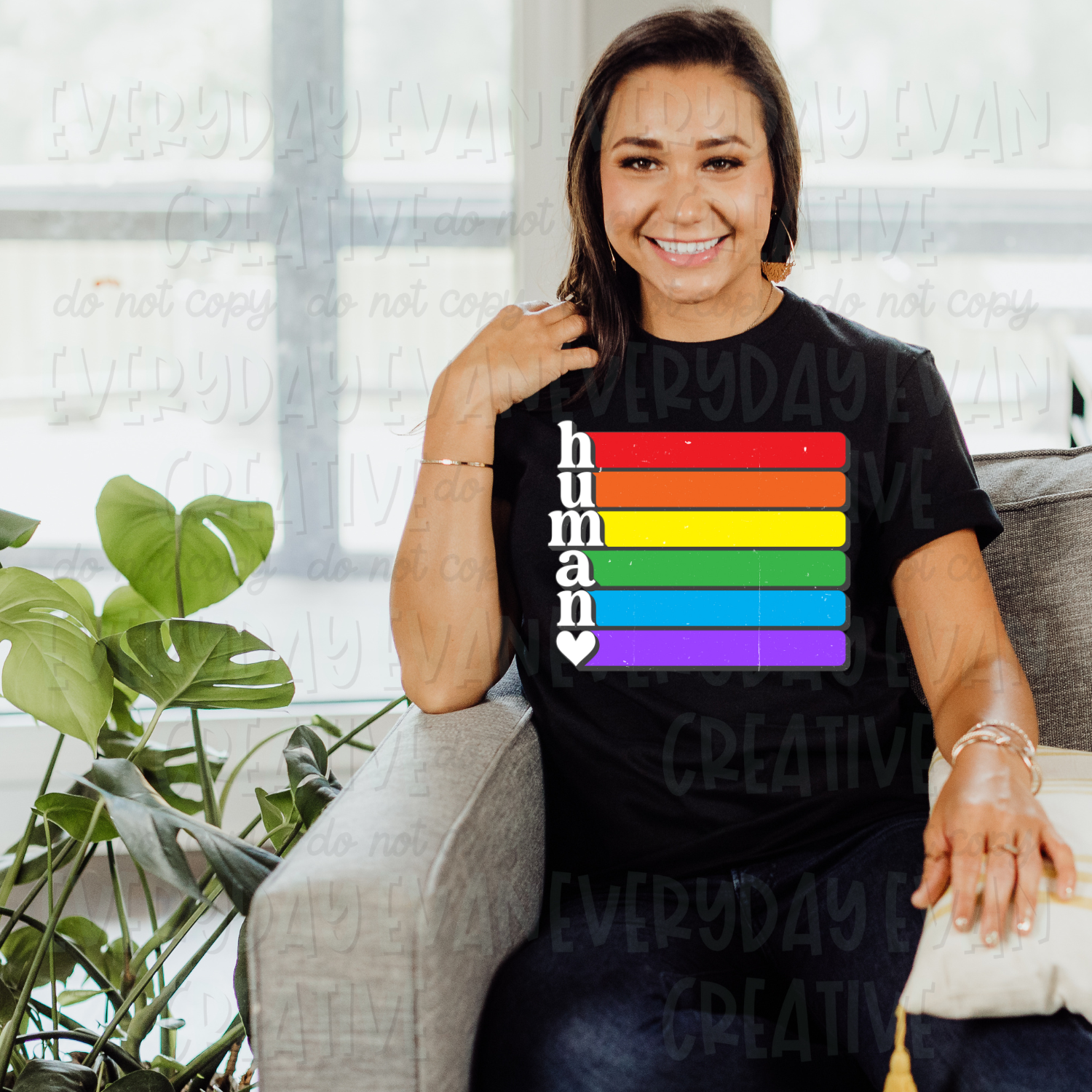 A brunette white woman sits on a couch modeling a black t-shirt. The design on the shirt has the word "human" vertically in white lowercase font. There is a white heart under the last letter. After each letter is a horizontal bar. The bars are in rainbow order from top to bottom: red, orange, yellow, green, blue, and purple.