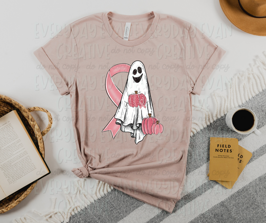 Image displays a pale pink t-shirt surrounded by miscellaneous desk items. The shirt's design includes a smiling sheet ghost holding a pink pumpkin, a large pink ribbon partially behind the ghost on the right side, and a couple of pink pumpkins beside the ghost on the lower left side.