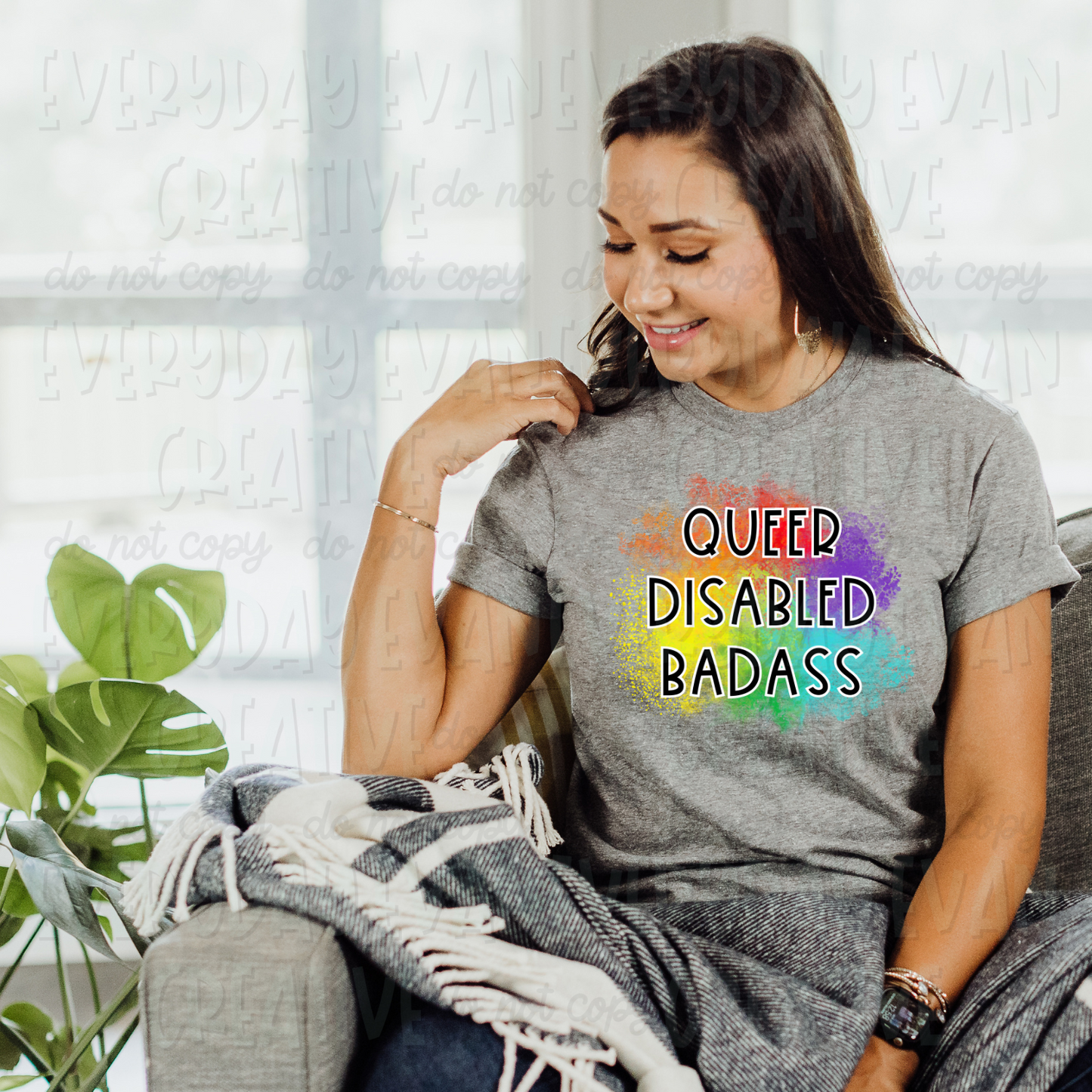 A brunette white woman sits on a couch modeling a sport gray t-shirt. The design on the shirt has a paint splatter background in rainbow colors. In the middle in thin black lettering outlined with white, it says "queer disabled badass" in all caps.