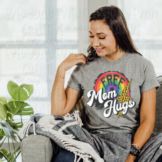 A brunette white woman sits on a couch modeling a sport gray t-shirt. The design on the shirt is a retro style design. It has a rainbow in the middle and a leopard print lightning bolt on the right. Inside the rainbow is the word "free" in all caps in dark gray. Underneath it says "Mom Hugs" in a white retro style cursive font. 