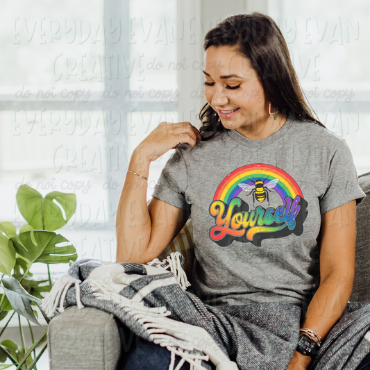 A brunette white woman sits on a couch modeling a sport gray t-shirt. The design on the shirt is a bright colored rainbow with a large bumble bee in the middle. Underneath it says "yourself" in a bold retro cursive font. The word is rainbow gradient in color with a dark gray offset.