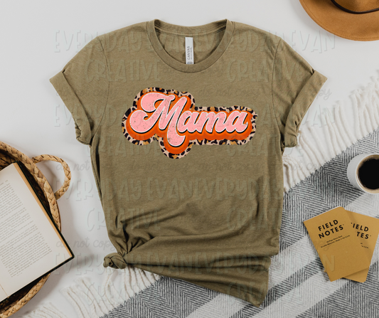 Image displays a t-shirt surrounded by miscellaneous desk items. The shirt's design includes a large script font that reads, "Mama." The text is surrounded and printed on top of different designs and colors. There is pink text, surrounded by a solid orange outline, and a cheetah print outline on the outermost edge hugging the letters.
