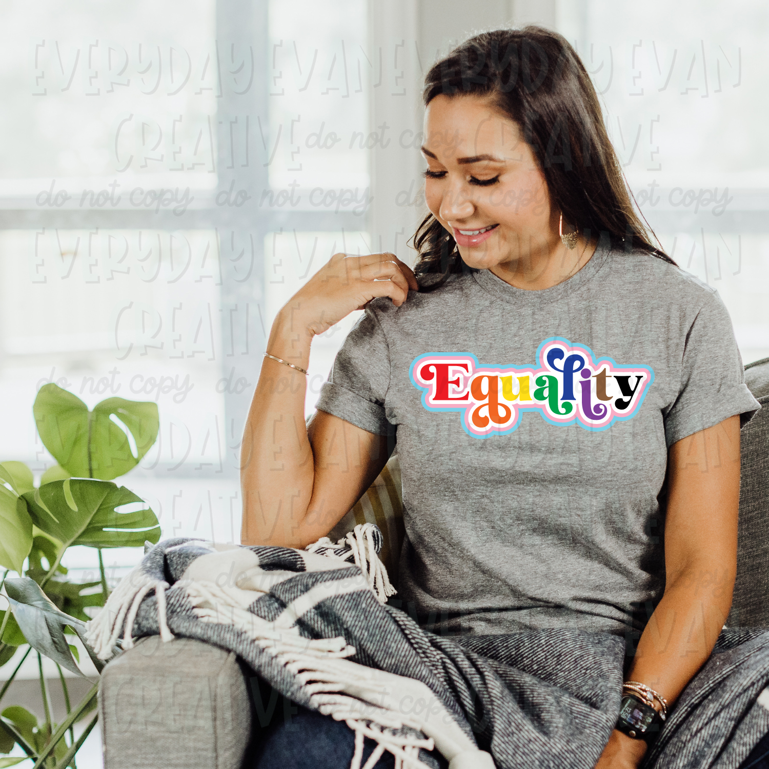 A brunette white woman sits on a couch modeling a sport gray t-shirt. The design on the shirt says "Equality." The letters in the word are in rainbow order: red, orange, yellow, green, blue, purple, brown, and black. There is an offset that is white, pink, and light blue. 