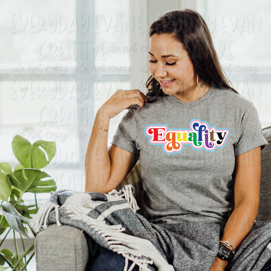 A brunette white woman sits on a couch modeling a sport gray t-shirt. The design on the shirt says "Equality." The letters in the word are in rainbow order: red, orange, yellow, green, blue, purple, brown, and black. There is an offset that is white, pink, and light blue. 