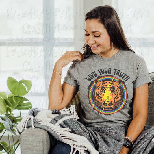 A brunette white woman sits on a couch modeling a sport gray t-shirt. The design on the shirt is a retro 80's style. It features a tiger's head with a lightning bolt through the middle surrounded by a circular rainbow with a dark gray offset. Above it says "live your truth" in a dark gray all caps retro font. 