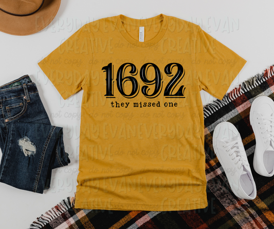 Image displays a t-shirt surrounded by miscellaneous clothing items. The shirt's design includes large western font that reads, "1692," and typewriter font text underneath that reads, "they missed one."