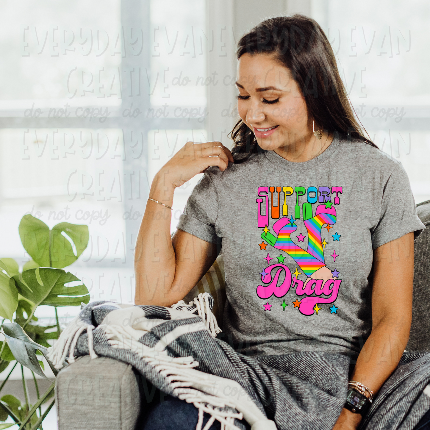 A brunette white woman sits on a couch modeling a sport gray t-shirt. The design on the shirt is brightly colored. It has rainbow striped go-go boots in the middle surrounded by rainbow starbursts. At the top it says "support" in a retro block font in rainbow colors. At the bottom is a bold retro cursive font in bright pink that says "Drag."