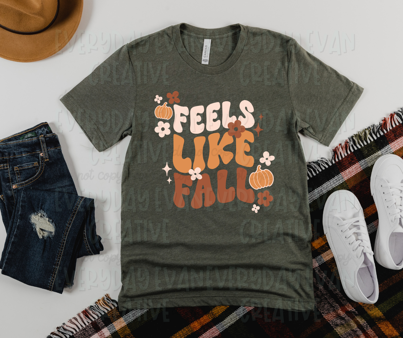 Image displays a t-shirt surrounded by miscellaneous clothing items. The shirt's design includes text that reads, "feels like fall" down the shirt in light pink, orange, and brown-red colors. The text is surrounded by small orange pumpkins, flowers, and starbursts of varying colors to match the text color scheme.