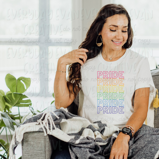 A brunette white woman is seated on a coach modeling a white t-shirt. The design on the t-shirt is the word "pride" in all caps in a hollow outline font. The word is repeated vertically six times in the following color order from top to bottom: red, orange, yellow, green, blue, purple.