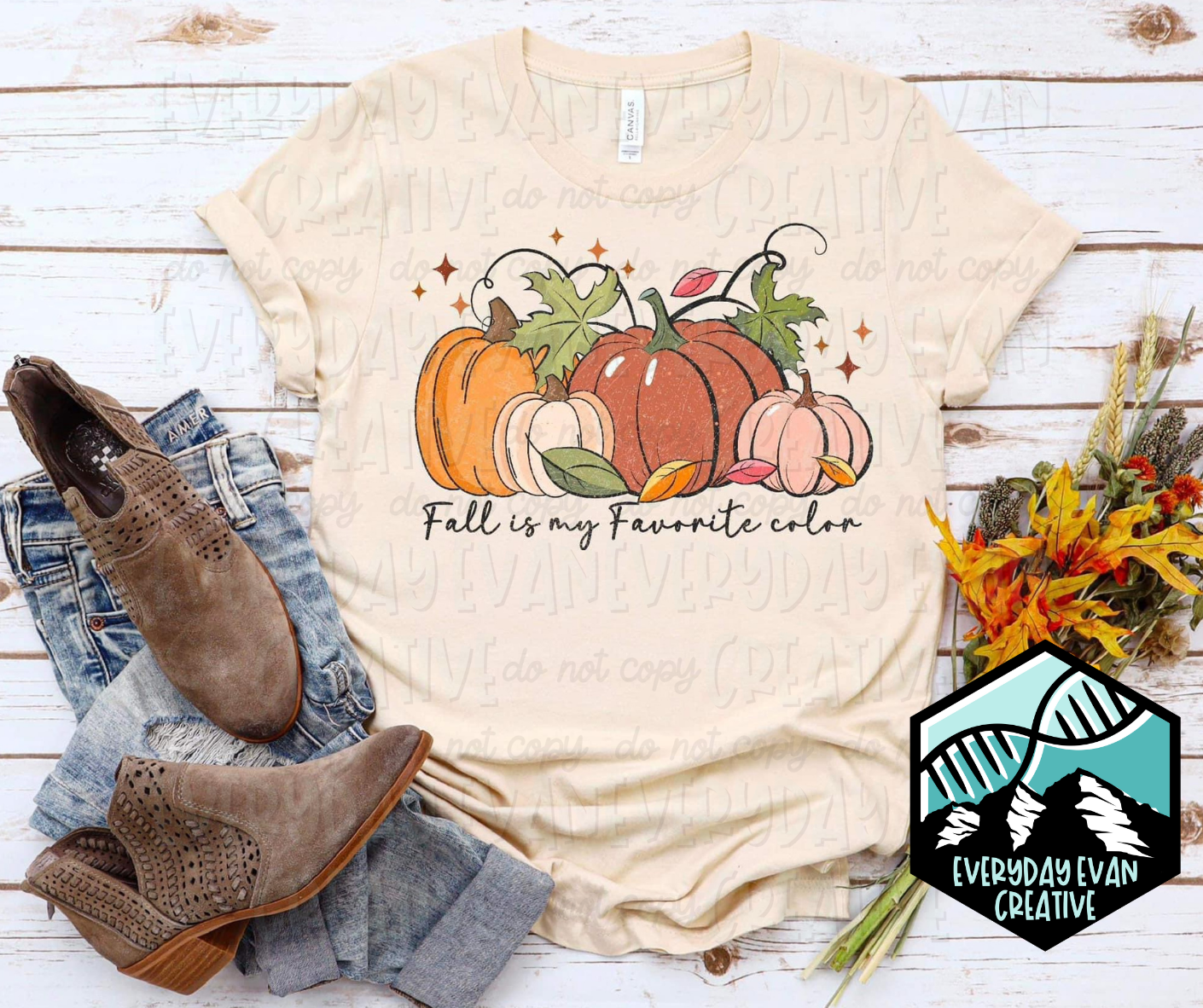 A flat lay of a cream colored t shirt with fall decor surrounding it. The design on the shirt has pumpkins of various colors and sizes and fall foliage with retro starbursts at the top. Underneath in script text it says "Fall is my Favorite color."