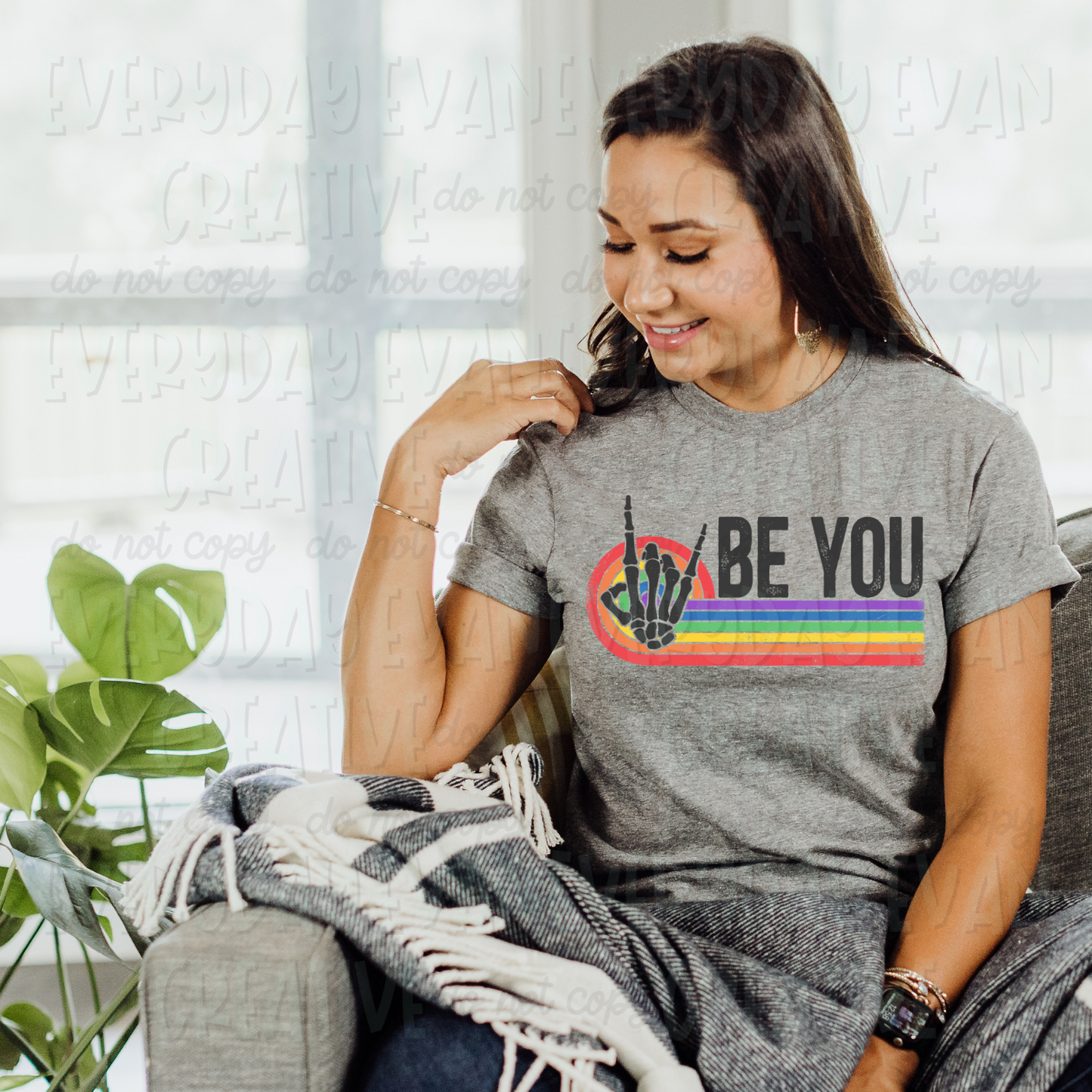 A brunette white woman sits on a couch modeling a sport gray t-shirt. The design on the shirt has a retro striped element in rainbow. There is a dark gray skeleton hand doing the rocker symbol with the index and pinky up. On top of the striped part of the design it says "be you" in all caps in a distressed dark gray block font.