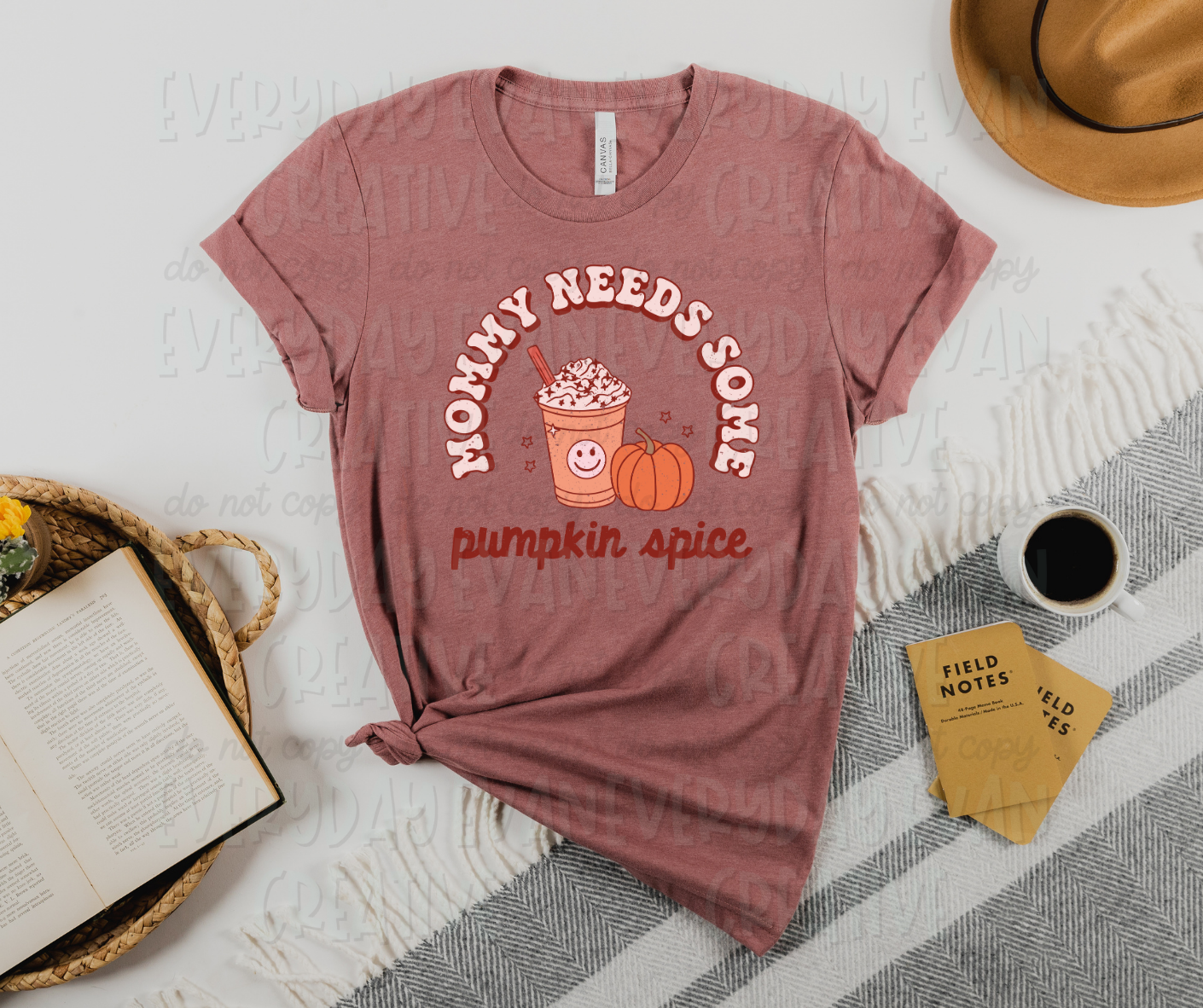 Image displays a t-shirt surrounded by miscellaneous desk items. The shirt's design has text in a semi-circle reading, "mommy needs some" and straight text across the bottom that reads "pumpkin spice." In the middle of the circle there is a coffee cup with a smile on it and an orange pumpkin. Small starbursts surround the cup and pumpkin.