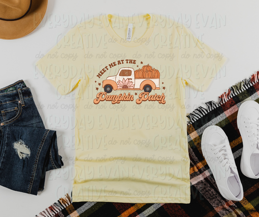 Image displays a t-shirt surrounded by miscellaneous desk items. The shirt's design includes a truck carrying orange pumpkins with a flower painted on the door. Above the truck, in a semi-circle of block text, it reads, "meet me at the," and below the truck, in script font, it reads "pumpkin patch." There are small starts surrounding the design.