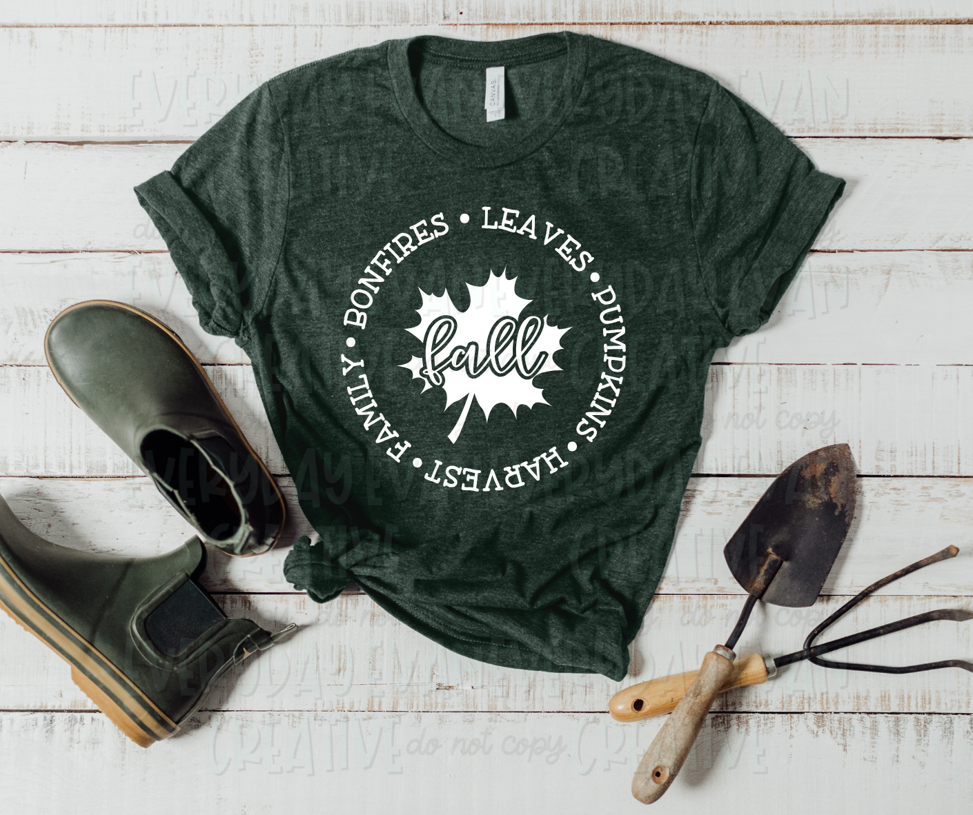 Image displays a t-shirt surrounded by boots and gardening tools. The shirt's design includes a white leaf with "fall" written in a script font in the middle. Around the leaf, a circle of words separated by dots surrounds the design, reading, "leaves, pumpkins, harvest, family, bonfires."