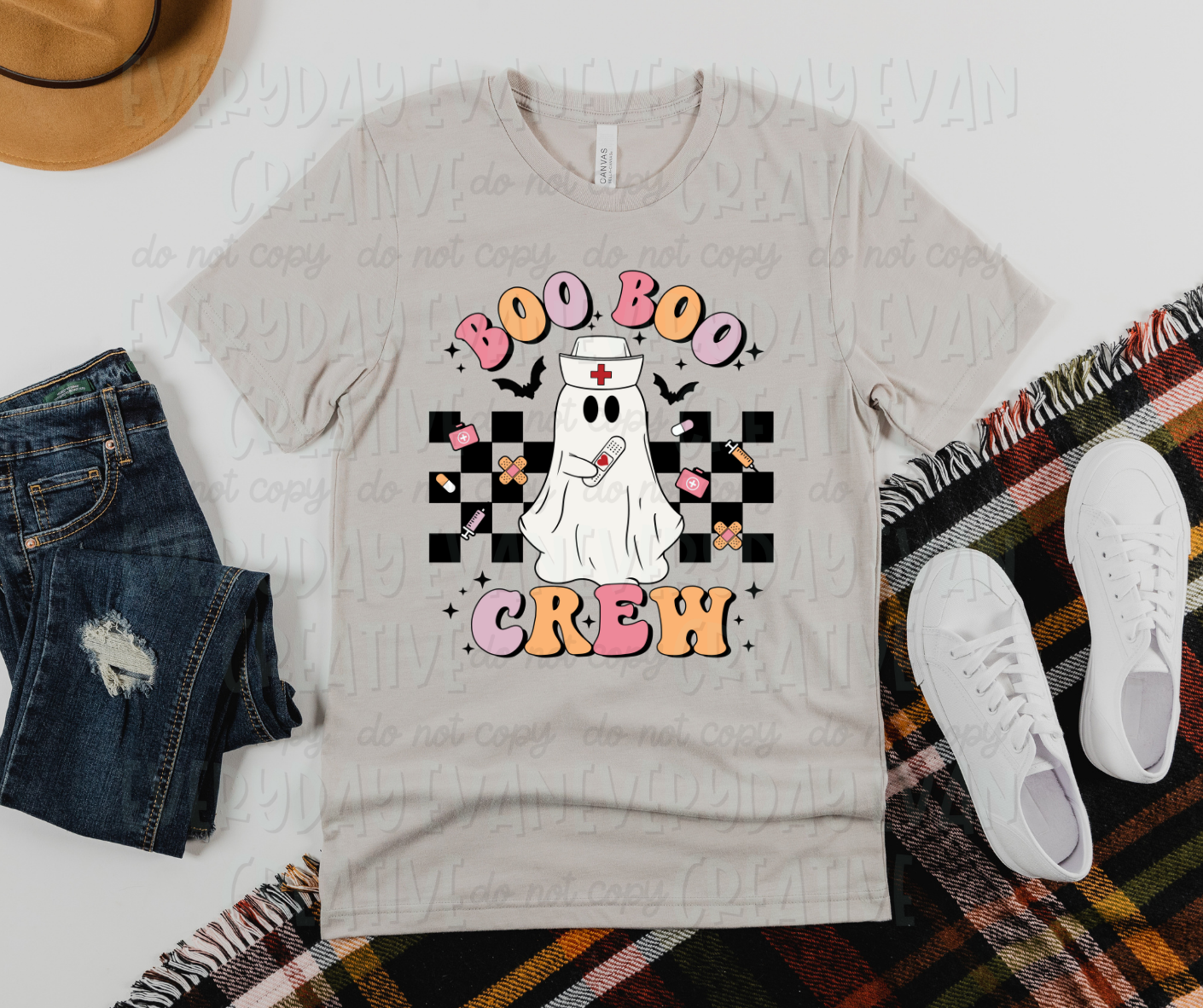 Image displays a t-shirt surrounded by miscellaneous clothing items. The shirt's design includes a checkered design covering the middle of the shirt with miscellaneous health items on top including band-aids, needles, and pills. In the middle of the checkered design, there is a sheet ghost with a nurse hat on and a band-aid with a heart on it in its hand. Bats and starbursts surround the design. Above the picture, in funky block font,  it reads, "Boo Boo," and on the bottom it reads "Crew."