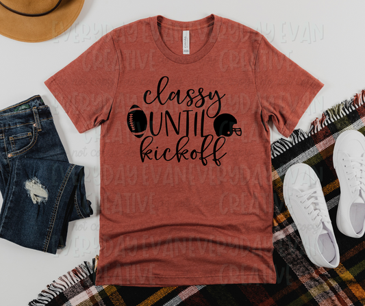 Image displays a t-shirt surrounded by miscellaneous clothing items. The shirt's design reads, "classy until kickoff" in both script and pencil font up and down the shirt with each word in its own row. Beside "until," (in the middle row) a football appears on the right and a football helmet appears on the left.