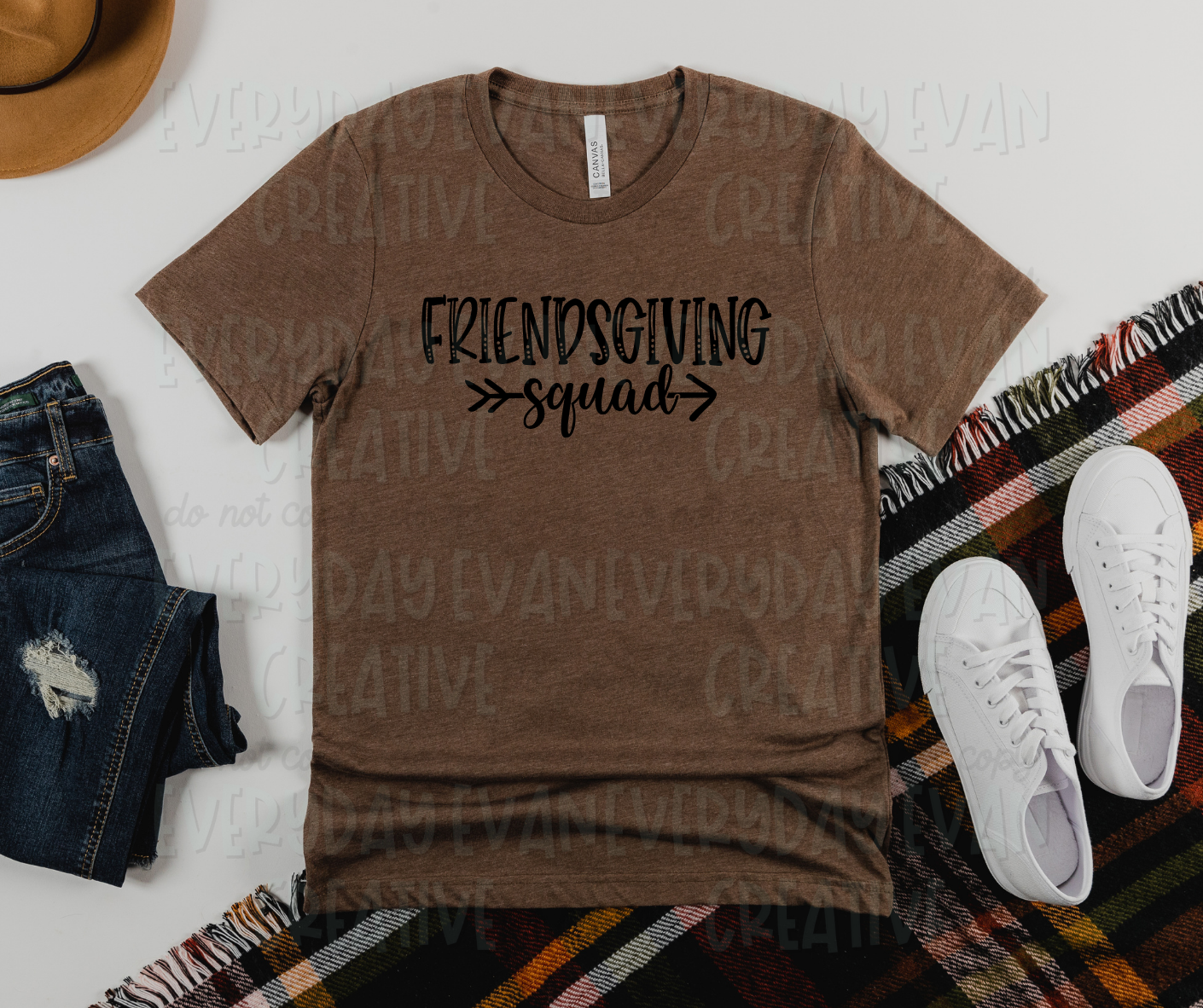 Image displays a t-shirt surrounded by miscellaneous clothing items. The shirt's design reads, "friendsgiving squad." "Friendsgiving" is in block pencil font, and "squad" is in script font. "Squad" appears in the middle of an arrow that points to the left 