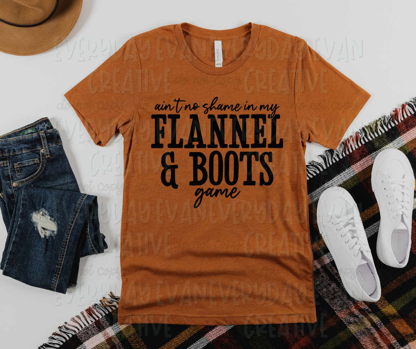 Image displays a t-shirt surrounded by miscellaneous clothing items. The shirt's design reads, "ain't no shame in my flannel and boots game." "And" appears as an ampersand. "Ain't no shame in my" and "game" appear in script font, while "flannel and boots" appears in thin block font.