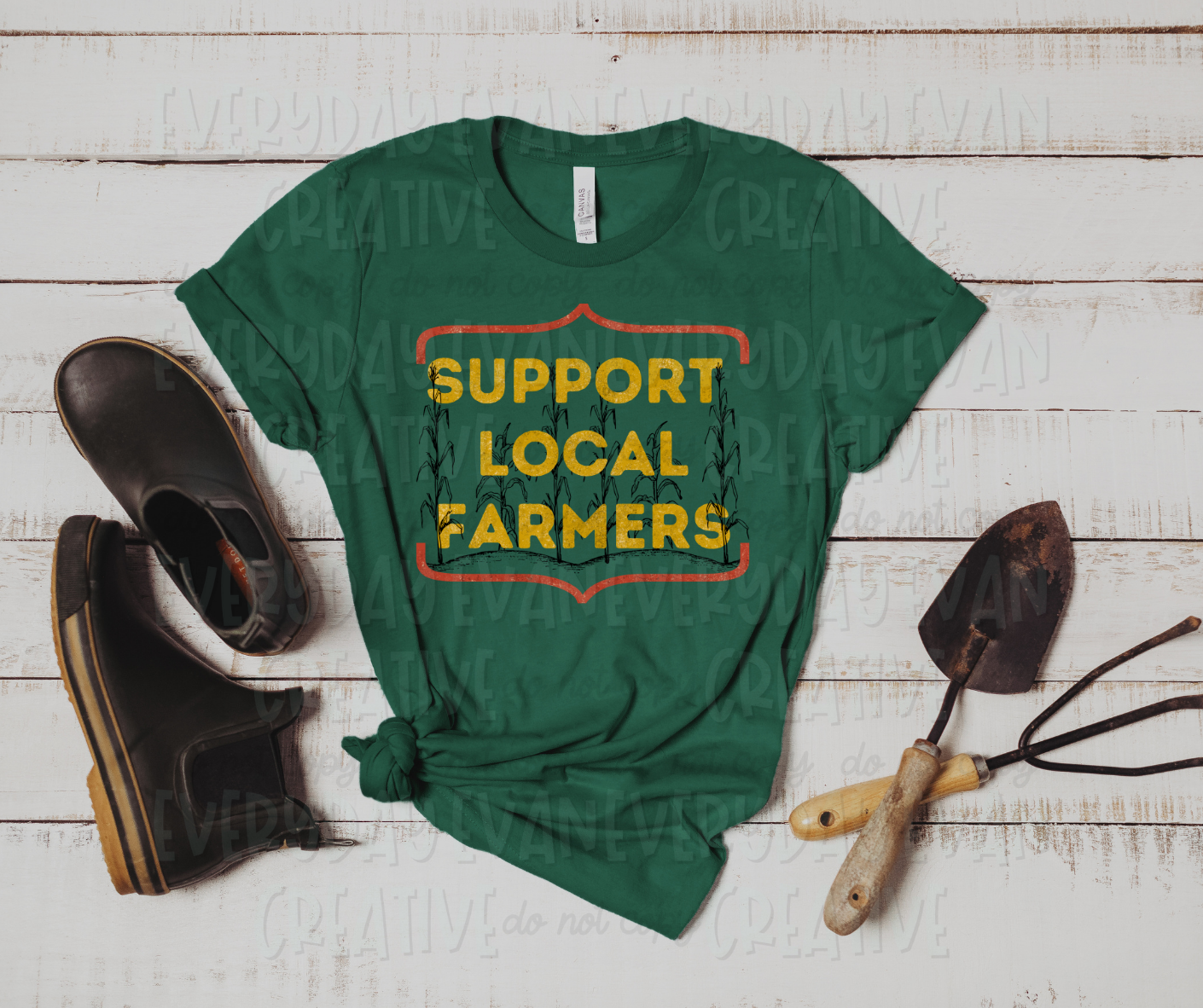 Image displays a t-shirt surrounded by boots and gardening equipment. The shirt's design includes yellow block font text that reads, "support local farmers" up and down the shirt with each word in it's own row. Behind the text, sketched corn stalks are growing from a ground. The design is encased by red braces on the top and bottom of the shirt.