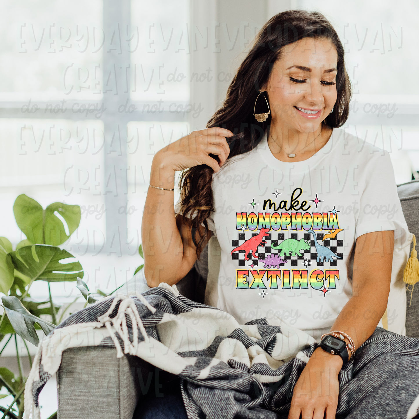 A brunette white woman sits on a couch modeling a white t-shirt. The design on the t-shirt is retro. In black cursive, it says "make." Underneath is a rainbow block font that says "homophobia." Underneath is a checkered background with multicolored dinosaur images. The bottom says "extinct" in the same rainbow block front as above. 