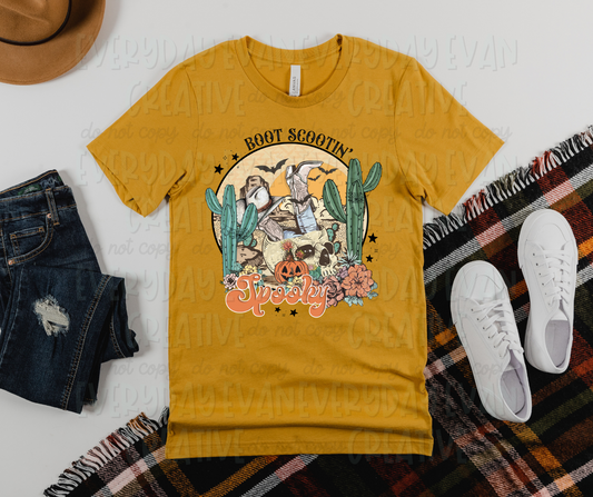 Image displays a shirt surrounded by fall themed clothing. The shirt has Halloween and country themed prints on it in a large circle, including a skull, cactus, boots, and a pumpkin in the circle. The circle has stars and flowers around it's edges to accent the main printed images. The circle is surrounded by text on the top and bottom. The top text reads, "boot scootin'" and the bottom text reads, "spooky."