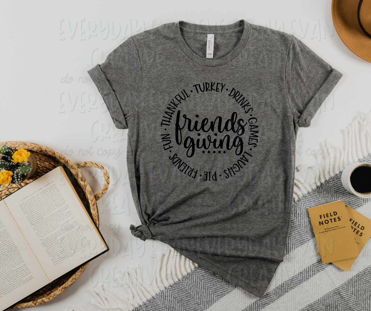Image displays a t-shirt surrounded by miscellaneous desk items. The shirt's design includes the text, "friendsgiving" written in a script font in the middle with dots underlining the text. Around the middle text, a circle of words separated by dots surrounds the design, reading, "turkey, drinks, games, laughs, pie, friends, fun, thankful."