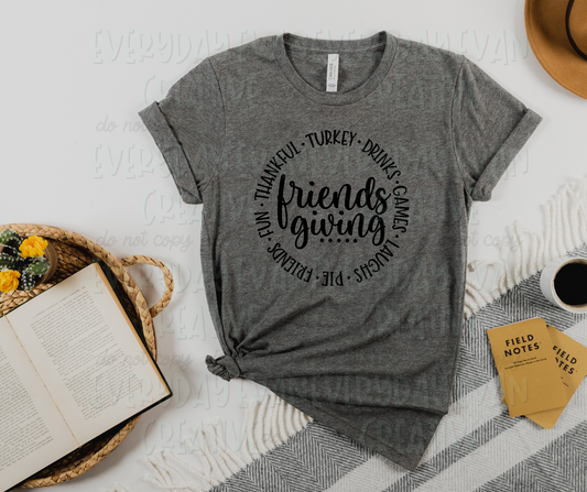 Image displays a t-shirt surrounded by miscellaneous desk items. The shirt's design includes the text, "friendsgiving" written in a script font in the middle with dots underlining the text. Around the middle text, a circle of words separated by dots surrounds the design, reading, "turkey, drinks, games, laughs, pie, friends, fun, thankful."