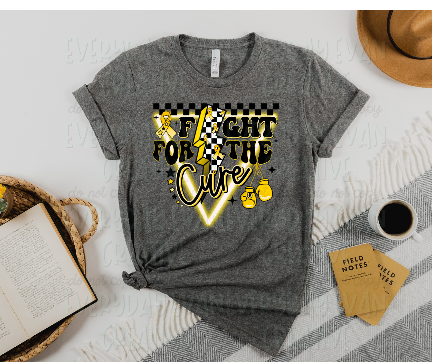 A flat lay of a t-shirt surrounded by miscellaneous desk items. The design features a neon upside down triangle with a checkered strip across the top. Black and yellow stars and starbursts, a pair of boxing gloves, and a yellow ribbon with stars in it surround the design. On top of the triangle, in different fonts, reads, "fight for the cure." The "i" in fight, is symbolized in the middle of the entire design with a large lightening bolt that has a checkered design and yellow ribbons printed in it.