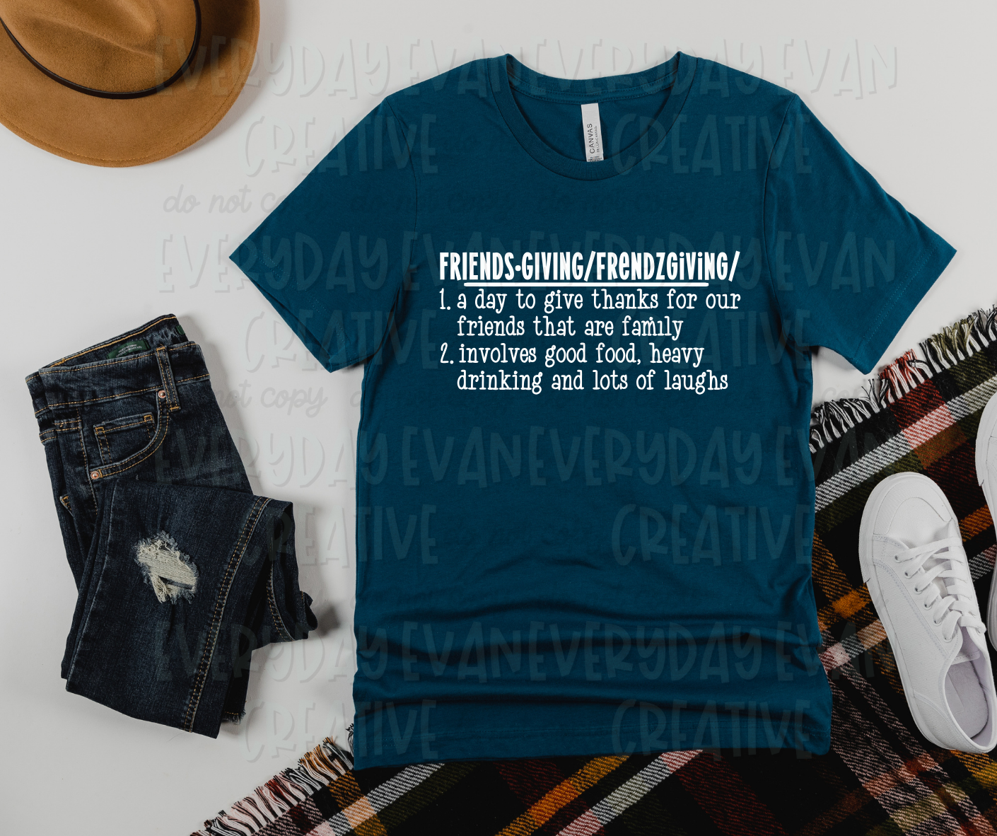 A flat lay of a t-shirt surrounded by miscellaneous clothing items. The shirt's design has a definition of friendsgiving. The text on the shirt reads, "friendsgiving/frendzgiving/1. a day to give thanks for our friends that are family. 2. involves good food, heaving drinking and lots of laughs."