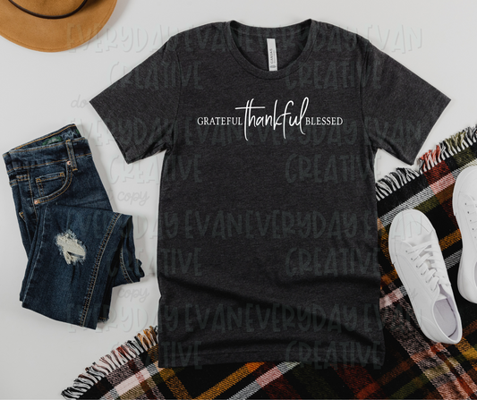 Image displays a t-shirt surrounded by miscellaneous clothing items. The shirt's design includes text across the top of the shirt that reads, "grateful thankful blessed" in serif and script font.