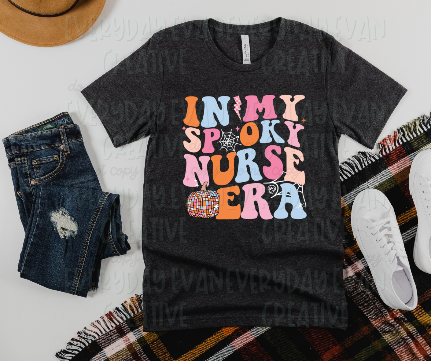 A flat lay of a t-shirt surrounded by miscellaneous clothing items. The shirt's design includes pink, orange, and light blue funky font text that reads, "in my spooky nurse era." Spiderwebs makeup one "o" in "spooky" and accent the shirt design as well. A disco-designed pumpkin, lightening blot, and starbursts are also in the design. 