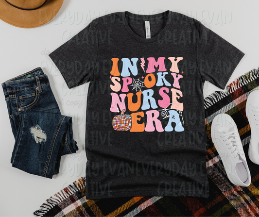 A flat lay of a t-shirt surrounded by miscellaneous clothing items. The shirt's design includes pink, orange, and light blue funky font text that reads, "in my spooky nurse era." Spiderwebs makeup one "o" in "spooky" and accent the shirt design as well. A disco-designed pumpkin, lightening blot, and starbursts are also in the design. 