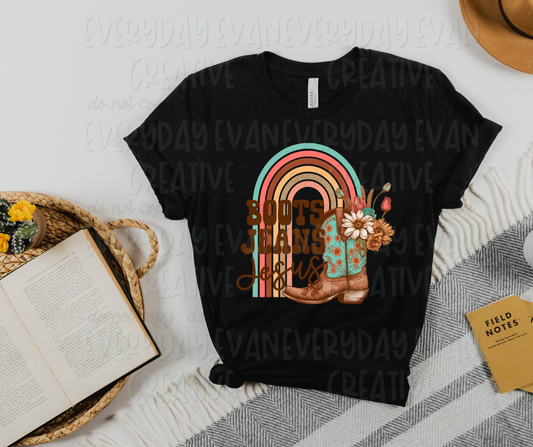 A flat lay of a t-shirt surrounded by miscellaneous desk items. The shirt design includes a rainbow ending in a cowboy boot. The boot has flowers and other foliage coming out of it along with the rainbow. On top of the design, western and script font reads, "boots jeans Jesus" on the rainbow side.