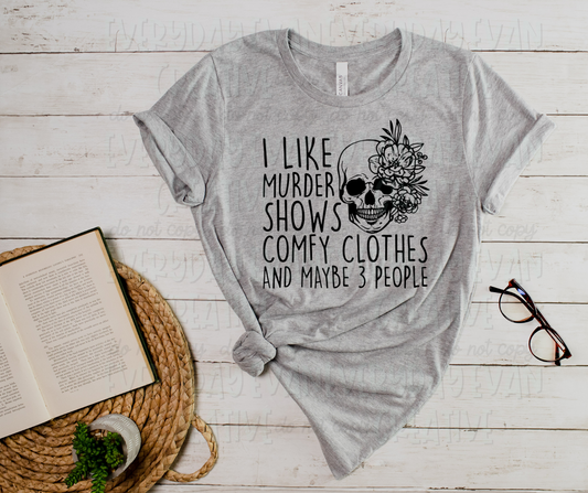 A flat lay of a t-shirt surrounded by miscellaneous desk items. The shirt's text reads, "I like murder shows comfy clothes and maybe three people," in a handwritten font. The text is wrapped around a skull with flowers in the top left-hand corner.