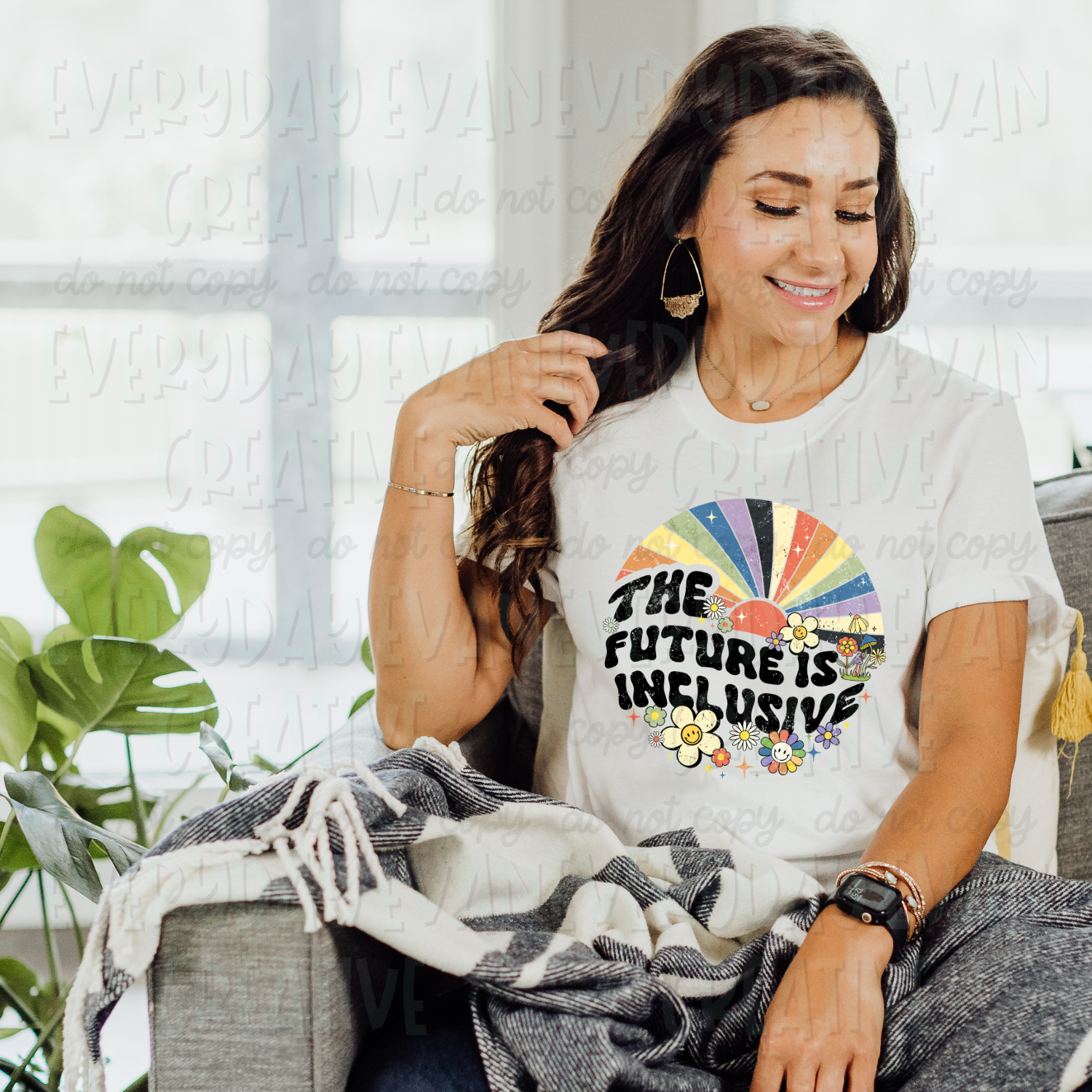 A brunette white woman is sitting on a couch modeling a white t-shirt. The design on the shirt is circular with a sunburst with rainbow rays at the top and groovy flowers at the bottom. In the middle in black retro distressed font, it says "the future is inclusive."