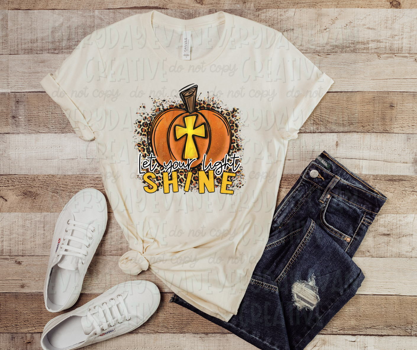 Image displays a shirt surrounded by dark jeans and white shoes. The shirt features an image of an orange pumpkin with a gold cross in the middle positioned on top of cheetah print which extends around the edges of the pumpkin. The pumpkin is positioned above text that reads, "let your light shine" in both script and block text font. "Shine," written in block letters, matches the same color as the cross in the pumpkin, while "let your light," written in script font, is black and white.