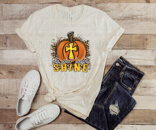 Image displays a shirt surrounded by dark jeans and white shoes. The shirt features an image of an orange pumpkin with a gold cross in the middle positioned on top of cheetah print which extends around the edges of the pumpkin. The pumpkin is positioned above text that reads, "let your light shine" in both script and block text font. "Shine," written in block letters, matches the same color as the cross in the pumpkin, while "let your light," written in script font, is black and white.