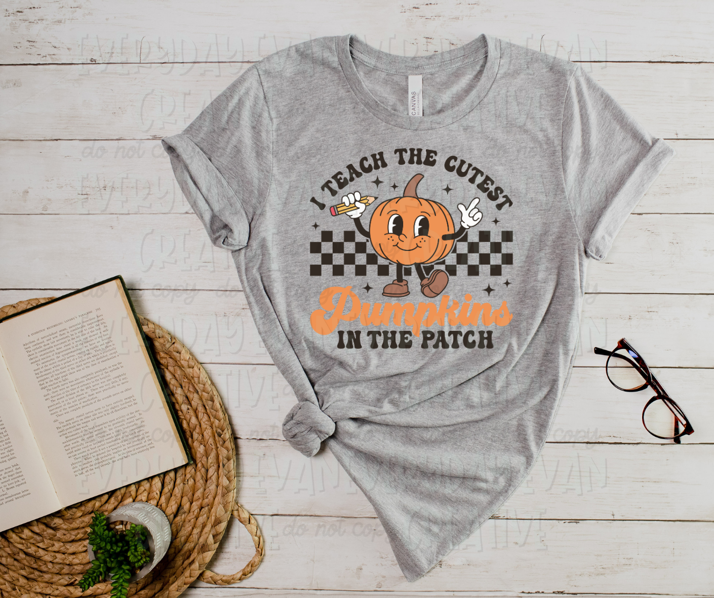 A flat lay of a t-shirt surrounded by miscellaneous desk items. The shirt's design includes a checkered design behind a personified pumpkin holding a pencil. Above the pumpkin, it text reads, "I teach the cutest" and below the design, it reads, "pumpkins in the patch." "Pumpkins" is in orange-colored script font, and the rest is in block font.