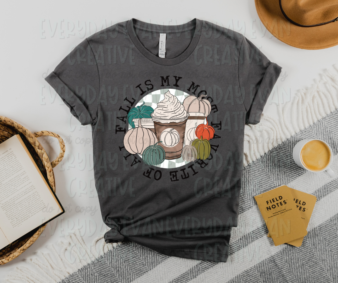 Image displays a t-shirt surrounded by miscellaneous desk items. The shirt's design includes a circle with a grey and white checkered background. In the circle, there are several coffee cups with pumpkin labels and several colored pumpkins surrounding them. The text surrounding the circle reads, "fall is my most favorite of all" flush with the design. 