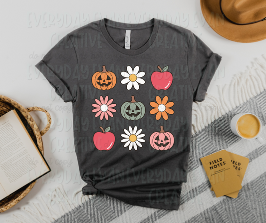 A flat lay with a t-shirt surrounded by miscellaneous desk items. The t-shirt design includes three rows of three images. Images include flowers of varying types, red apples, and three Halloween-themed orange, pink, and pale green pumpkins.