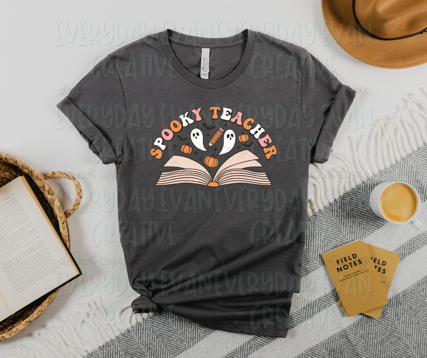 Image displays a t-shirt surrounded by miscellaneous desk items. The shirt's design includes an open book with ghosts, pumpkins, bats, and starbursts above the pages. Above the design, in funky block font, it reads, "spooky teacher" in alternating orange, white, and pink letters.