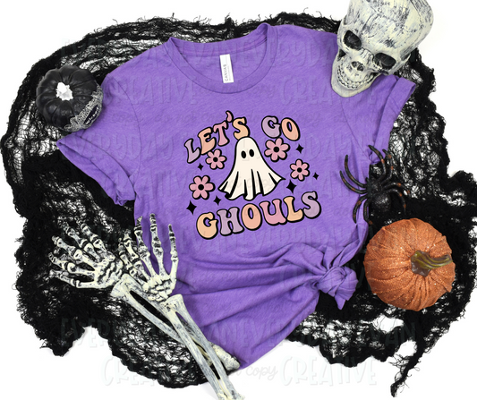 A flat lay of a t-shirt surrounded by Halloween-themed items. The shirt's design includes a sheet ghost with flowers and starbursts surrounding it. The text above and below the design, in funky block font, reads "let's go ghouls."